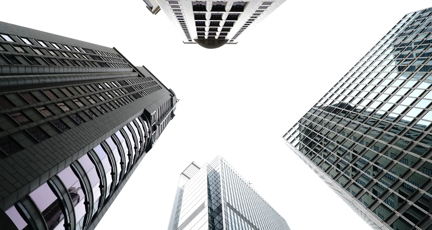 Looking straight up between four high-rise office towers, their glass and stone facades converging on a bright white sky.