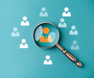 A magnifying glass over a teal background enlarges one orange figure among a scattering of white person icons.