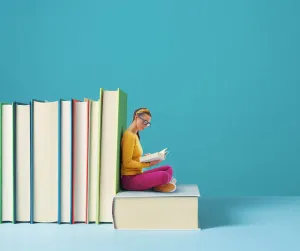A woman in a yellow sweater reads, seated on an oversized book beside a row of giant books against a teal wall.