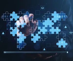 A hand fits glowing blue puzzle pieces together above a tablet, surrounded by linked people icons.